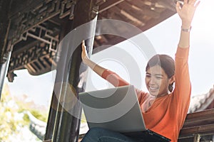 Happy young asian woman be smile working with her laptop and hand up on a bench in the park outdoors on vacation time