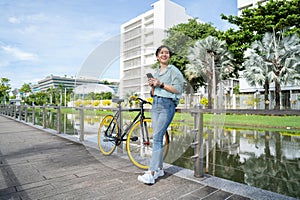 Happy young Asian woman in casual clothes standing near bicycle on sidewalk in city park. She is using a smartphone