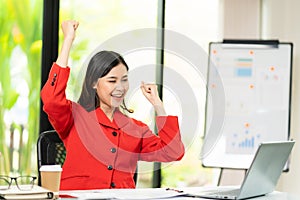 Happy young Asian woman celebrates success with arms raised in front of laptop in office, finance business concept