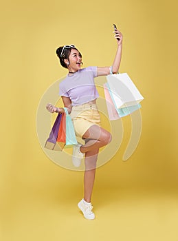 Happy young asian woman celebrating with mobile phone and holding shopping bags isolated over yellow background