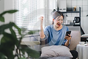 Happy young asian woman celebrating success with arms raised in front of a mobile phone, fists clenched.