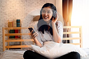 Happy young Asian woman in earphones singing and listening to music while sitting on bed and holding mobile phone at