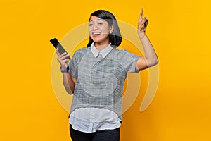 Happy young Asian woman holding mobile phone and pointing finger to copy space on yellow background