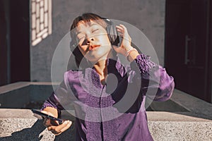 Happy young asian woman listening to music with headphones via smartphone and having fun while sitting side the street.