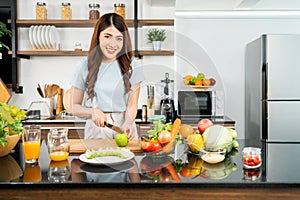 A happy young Asian woman looking at the camera preparing a healthy salad with vegetables on a cutting board in the home kitchen.