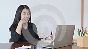 Young asian woman looking at laptop computer screen and making notes on notebook.