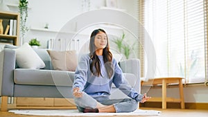 Happy young Asian woman practicing yoga and meditation at home sitting on floor in living room in lotus position