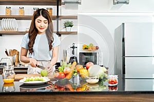 A happy young Asian woman preparing a healthy salad with fresh vegetables on a cutting board in the home kitchen. Food cooking.