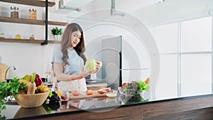 A happy young Asian woman preparing a healthy salad with vegetables on a cutting board on the home kitchen counter.