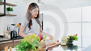 A happy young Asian woman preparing a healthy salad with vegetables on a cutting board on the home kitchen counter.