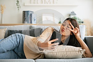 Happy young asian woman relax on comfortable couch at home texting messaging on smartphone