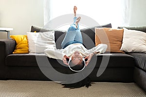 Happy young Asian woman relaxing at home, lying on the couch upside down, listening to music with headphones.