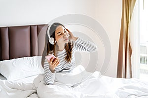 Happy young asian woman relaxing and using headphones to listen to music from smart phone in bedroom