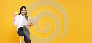 Happy young Asian woman showing credit card and using computer online shopping isolated over yellow background