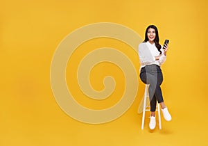Happy young Asian woman showing mobile phone. While her sitting on white chair and looking on yellow copy space background