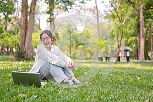 A happy young Asian woman sits on the grass in a park, smiling while looking at her laptop