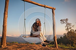 Happy young asian woman sitting and smiling at wooden swing on top of mountain at the evening