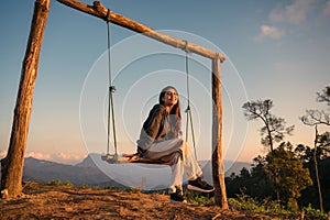 Happy young asian woman sitting and smiling at wooden swing on top of mountain at the evening
