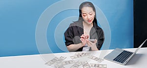 Happy young asian woman smile while hand holding smart phone and a lot of USD currency banknotes on work table with notebook