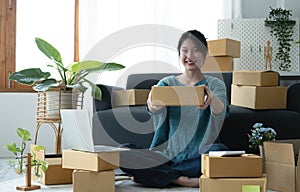 Happy young asian woman startup small business freelance holding parcel box and computer laptop and sitting on floor, Online