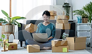Happy young asian woman startup small business freelance holding parcel box and computer laptop and sitting on floor, Online