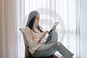 Happy young Asian woman using tablet while sitting at condominium. Technology and lifestyle, leisure activities on weekend concept
