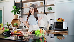 A happy young Asian woman waving hands while preparing a healthy salad with vegetables on the home kitchen counter.