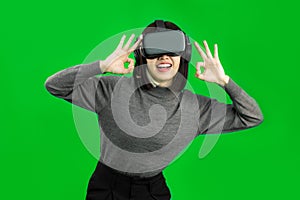 Happy Young Asian woman wearing a gray shirt and a VR headset with OK sign, greenscreen
