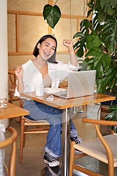 Happy young asian woman winning on laptop, receive good news, achieve goal at work, triumphing and smiling pleased