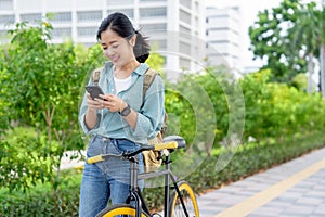 Happy young Asian woman in work clothes using smartphone with bicycle in outdoor park. Eco-friendly concept