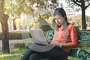 Happy young asian woman working with her laptop on a bench in the park outdoors on vacation time