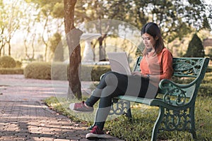 Happy young asian woman working with her laptop on a bench in the park outdoors on vacation time