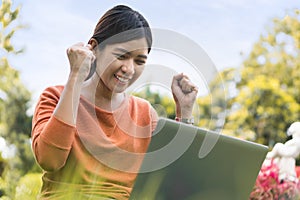 Happy young asian woman working with her laptop on a bench in the park outdoors on vacation time