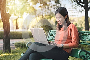 Happy young asian woman working with her laptop on a bench in the park outdoors on vacation time