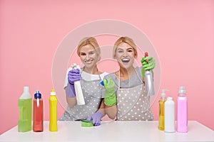Happy young attractive blonde cleaning ladies dressed in uniform raising hands with spray bottles and smiling gladly at camera,