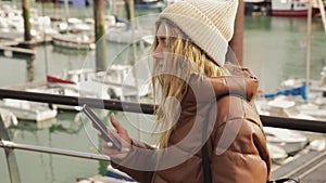 Happy young attractive stylish girl blogger is walking by the port and looking at her cell phone at sunset with boats on