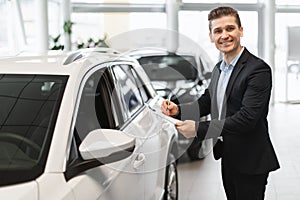Happy young auto insurance agent standing near new car, checking it, taking notes, working at automobile dealership