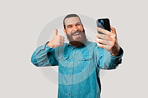 Happy young bearded man is taking a selfie with his phone shows a thumb up.