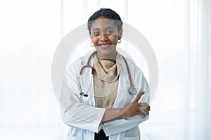 Happy young beautiful African American female doctor in white medical coat with stethoscope standing with crossed arms and smiling