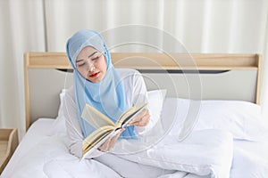 Happy young beautiful asian muslim woman in sleepwear sitting, reading book on bed. Cheerful and cute girl resting in bedroom and