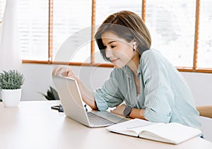 Happy young beautiful Asian woman using laptop working remote and wireless earphones sitting at table in home office.Remote work,E