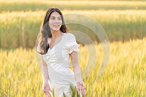 Happy young beautiful Asian woman in white dress standing in the barley field enjoying her time outdoor alone during sunset