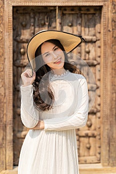 Happy young beautiful attractive asian woman in white dress and straw hat smiling at camera while standing ouside a rustic wooden