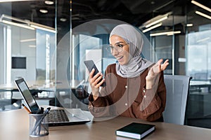 Happy young beautiful muslim woman in hijab working in office at desktop and looking enthusiastically at phone screen