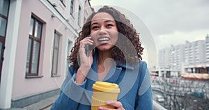 Happy young beautiful successful mixed race business woman talks on the phone smiling, holds coffee cup in city street.