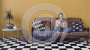 Happy young and beautiful woman playing a game on her smart phone, sitting on a couch at home in bright stylish room