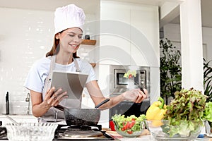 Happy young beautiful woman wearing apron and chef hat in kitchen, lovely girl holding digital tablet for searching tasty healthy