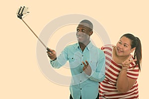 Happy young black African man and young fat Asian woman smiling while giving peace sign and taking selfie picture with