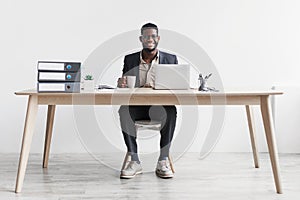 Happy young black businessman with coffee working online on laptop, sitting at desk and smiling at camera