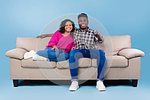 Happy young black couple with remote control sitting on sofa and watching TV over blue studio background, full length